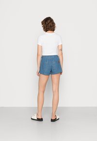 Kvinna med kort lockigt hår står vänd bort, iklädd en vit kortärmad topp, blå denimshorts, vita sandaler och en tatuering på underarmen.