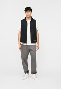 Jeune homme debout portant une veste noire zippée sur un t-shirt blanc, un pantalon de survêtement gris et des baskets blanches, sur un fond clair uni.