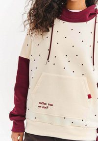 Kremowa bluza z kapturem z bordowymi akcentami i wzorem w kropki. Posiada przednią kieszeń oraz napis "coffee, tea, or me?" w kolorze bordowym.