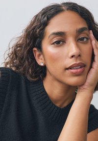 Schwarzer, gerippter Strickpullover mit hohem Ausschnitt. Das Haar ist lockig mit glänzender Textur. Das Modell trägt kleine, runde Steckerohrringe.