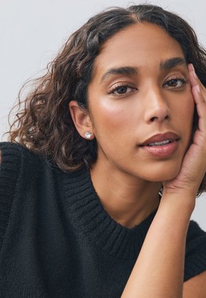 Black ribbed knit sweater featuring a high neckline. Hair is curly with a glossy texture. Model wears small, round stud earrings.