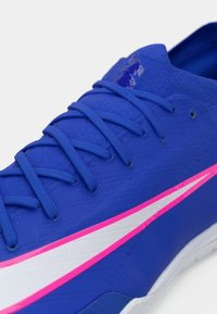 Μπλε αθλητικό παπούτσι με υφή στο πάνω μέρος, διαθέτει ροζ και λευκές λεπτομέρειες. Περιλαμβάνει κορδόνια και μια λείεια σόλα που είναι ορατή στην εικόνα.