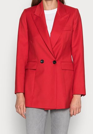 Femme portant un blazer rouge vif à double boutonnage avec des boutons noirs sur une chemise blanche et un pantalon gris clair.