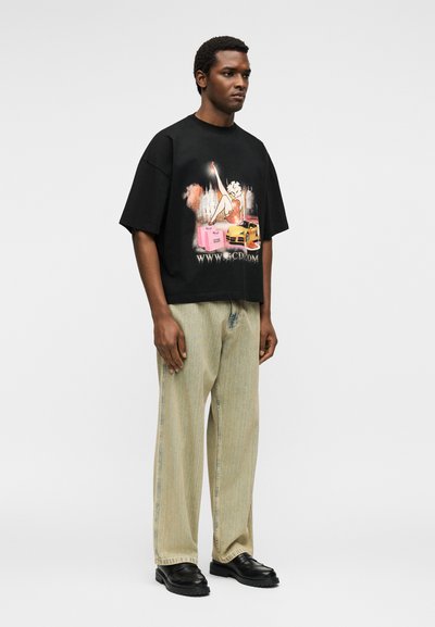 Άνδρας στέκεται φορώντας μαύρο μεγάλο T-shirt με πολύχρωμο καρτουνίστικο σχέδιο, μπεζ φαρδιά τζιν και μαύρα chunky παπούτσια σε λευκό φόντο.