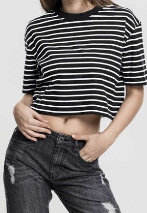 Person trägt ein schwarz-weiß gestreiftes, kurz geschnittenes T-Shirt und hoch taillierte, schwarze Jeans mit Used-Effekten, mit einer Hand in der Tasche.