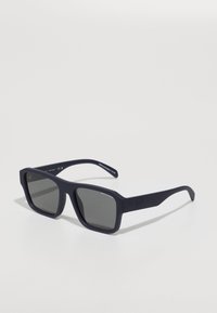 Occhiali da sole blu navy con finitura opaca, lenti quadrate e montatura spessa. Presentano un brand discreto sulla tempia.