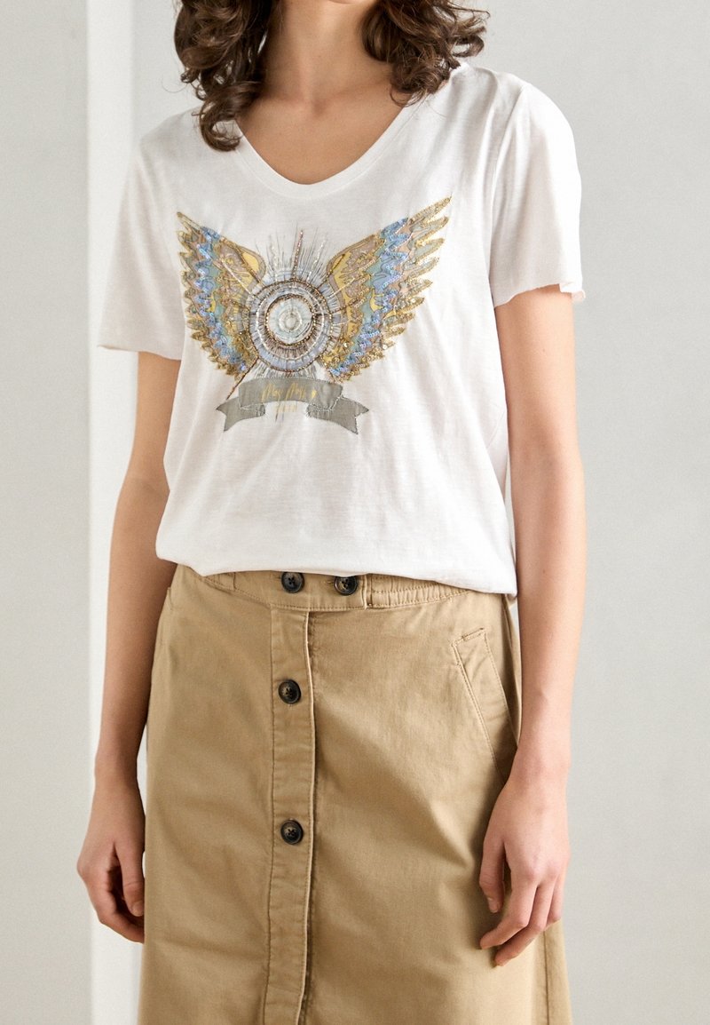 Bílý tričko s krátkým rukávem s dekorativním motivem modrých a zlatých křídel, který je spárován s béžovou sukní na knoflíky.