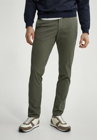 Uomo che indossa pantaloni slim-fit verde oliva con sneaker bianche e marroni, in piedi contro uno sfondo chiaro uniforme.