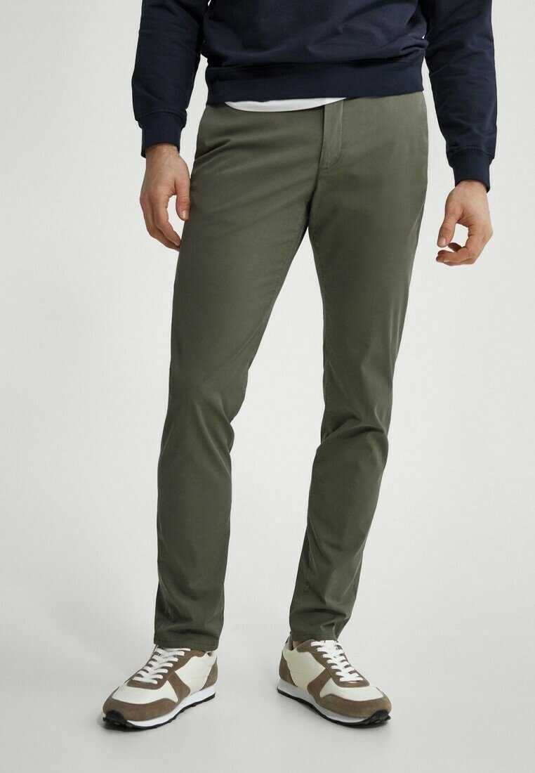 Uomo che indossa pantaloni slim-fit verde oliva con sneaker bianche e marroni, in piedi contro uno sfondo chiaro uniforme.