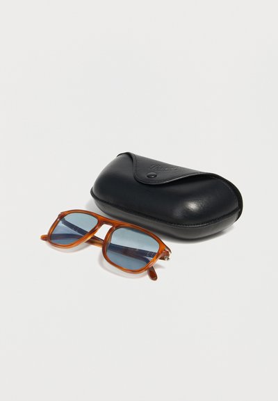 Naočale s smeđim okvirom i plavim lećama pored zatvorenog crnog kože kućišta s kopčom na bijeloj površini.