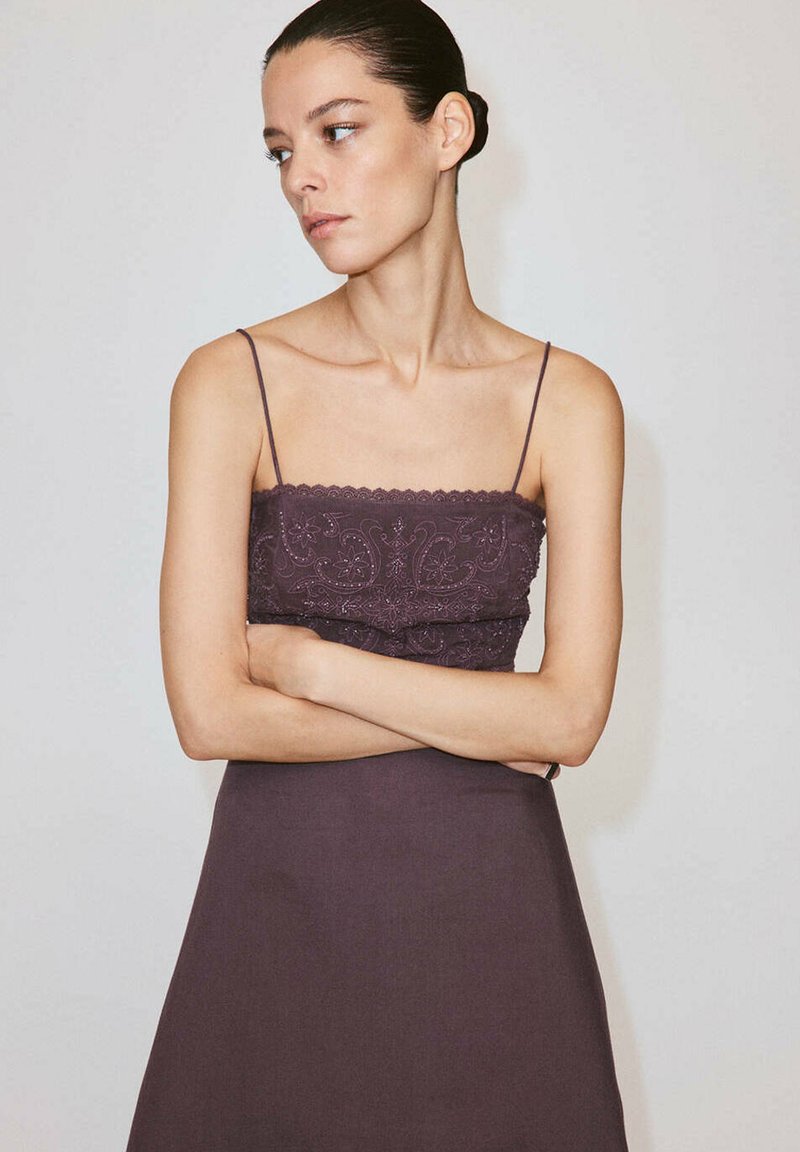 Femme aux cheveux foncés attachés, portant une robe sans manches violet foncé avec fines bretelles et détails en dentelle, bras croisés, regardant sur le côté.