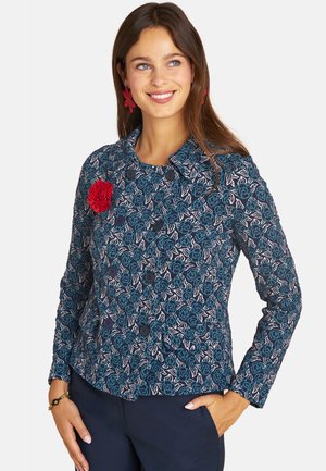 Frau lächelnd, trägt eine blaue Jacke mit floralem Muster und schwarzen Knöpfen, rote Blumenbrosche, rote Ohrringe und marineblaue Hose, Hand in der Tasche.