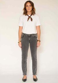 Femme debout, portant une chemise blanche �à manches courtes, un jean gris foncé, des ballerines noires cloutées, un foulard marron autour du cou et des bracelets dorés.