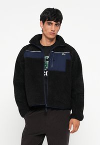 Schwarz gefütterte Jacke mit einem marineblauen Einsatz, Reißverschlusstaschen, hohem Kragen und hellblauen Akzenten. Verfügt über ein strukturiertes, weiches Material.