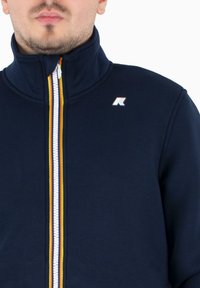 Giacca a zip blu navy con colletto alto, caratterizzata da una zip a righe multicolori e un piccolo logo sul petto. Tessuto liscio, design casual.
