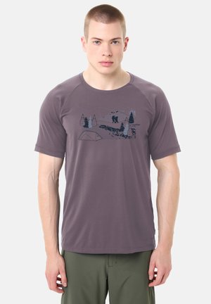 Homme portant un t-shirt violet avec un graphique de forêt sombre et de randonnée, associé à un pantalon vert, debout devant un fond uni.