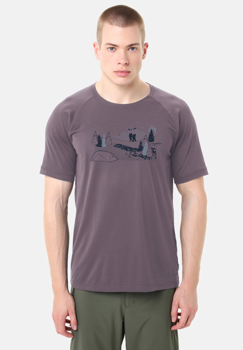 Homme portant un t-shirt violet avec un graphique de forêt sombre et de randonnée, associé à un pantalon vert, debout devant un fond uni.