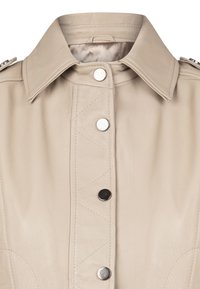 Giacca in pelle beige con colletto strutturato, bottoni a pressione in argento, dettagli trapuntati e una texture liscia. Nessun motivo visibile o accessori in metallo.