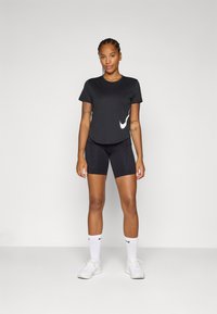 Une femme se tient avec confiance, portant un t-shirt noir Nike et un short, associés à des chaussettes blanches et des baskets, sur un fond uni.
