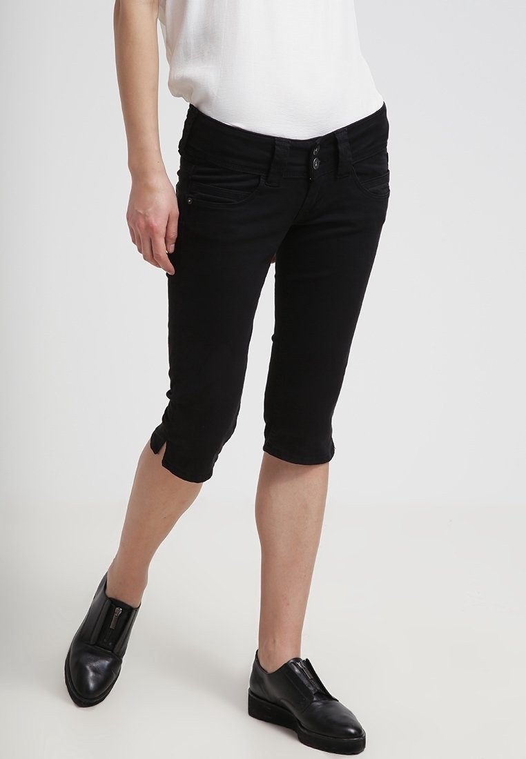 Personne portant un pantalon noir ajusté mi-mollet, un haut blanc et des chaussures en cuir noires sans lacets, debout sur un fond clair.