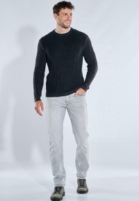 Dunkelgrauer, gerippter Pullover mit rundem Ausschnitt, kombiniert mit hellgrauen Jeans und grünlich-braunen Schuhen, stehend vor hellem Hintergrund.
