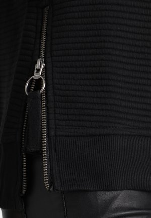 Close-up van een zwart geribd stoffen jack met zilveren rits en ringvormige trekker, gedragen over zwarte leren broek.