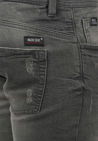 Jeans de denim gris oscuro con una textura desgastada, que presentan un parche con el logo, múltiples bolsillos y un cierre de botón.