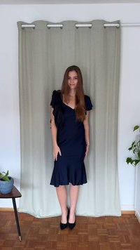 Mélykék, testhez simuló ruha V-nyakkivágással és fodros fekete részletekkel a vállakon. Bővülő szegély. Fekete, hegyes orrú magassarkúval viselve.