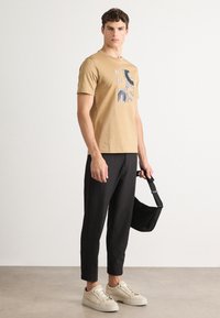 Beige bomulls T-skjorte med grafisk trykk, paret med svarte tettsittende bukser og kremfargede joggesko. Modellen holder en svart veske med stropp.