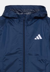 Veste légère bleu marine avec capuche, fermeture éclair, accents élastiques et logo à trois barres blanches sur la poitrine gauche. Texture lisse.