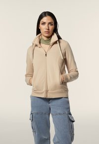 Felpa con zip color beige, dotata di pannelli in pile testurizzato, tasche laterali e cappuccio con cordino. Il modello indossa un sottogonna verde chiaro e pantaloni cargo blu chiaro.