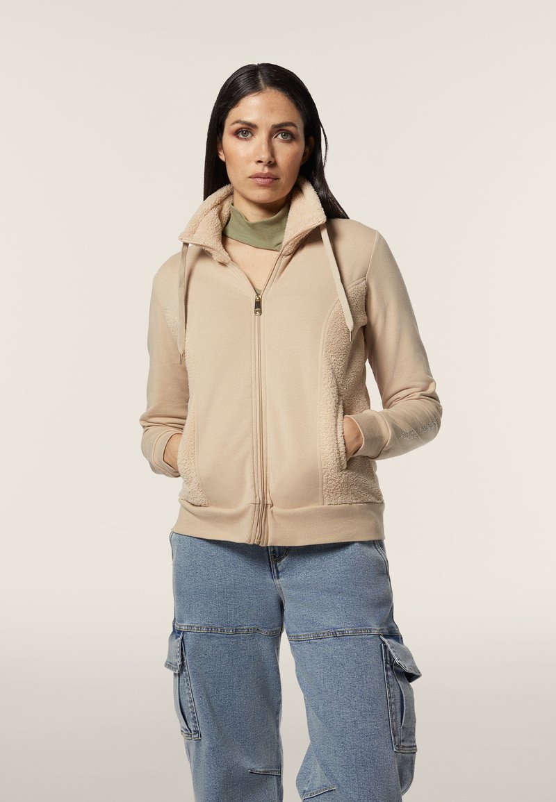 Felpa con zip color beige, dotata di pannelli in pile testurizzato, tasche laterali e cappuccio con cordino. Il modello indossa un sottogonna verde chiaro e pantaloni cargo blu chiaro.