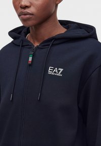 Άτομο που φοράει ένα σκούρο μπλε φούτερ Emporio Armani EA7 με φερμουάρ και κουκούλα, με τα χρώματα της ιταλικής σημαίας στην τσέπη του φερμουάρ.