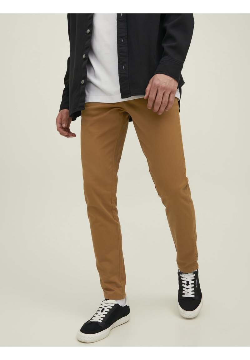 Homme portant un pantalon slim marron, des baskets noires avec des lacets blancs, des chaussettes blanches, un t-shirt blanc et une chemise noire boutonnée.