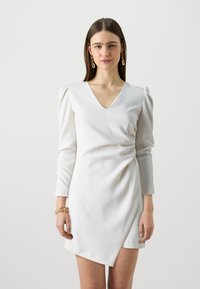 Fehér, hosszú ujjú, V-nyakú, ráncolt vállrészű csomagolóruha. Simító anyagból készült, enyhén aszimmetrikus szegéllyel.