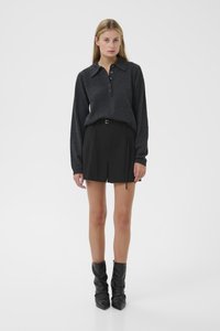 Mujer vestida con una polo de manga larga gris oscuro, pantalones cortos negros con cinturón y botas puntiagudas negras y holgadas, de pie contra un fondo blanco.