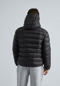 Sort pufferjakke med hætte, der har vandrette quiltede sektioner, glat tekstur og elastiske manchetter. Bæres med grå sweatpants.