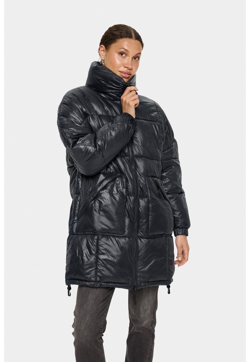 Pūkains melns mētelis ar augstu apkakli un rāvējslēdzēja aizdari. Aprīkots ar divām priekšējām kabatām un elastīgām aprocēm. Spīdīgs audums ar šūtu rakstu.