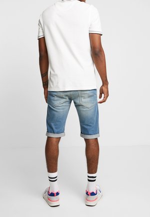 T-shirt blanche avec bord noir, accompagnée de shorts en denim bleu clair avec un ourlet roulé. Baskets blanches avec accents roses et bleus.