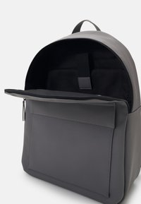 Sac à dos gris avec un extérieur lisse, un haut arrondi, une poche zippée à l'avant et un intérieur noir. Comprend une poignée supérieure et un design minimaliste.
