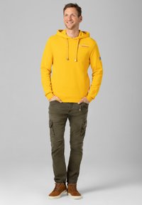 Gelbes Kapuzensweatshirt mit Kordelzügen und Kängurutasche; kombiniert mit olivgrünen Cargo-Hosen und braunen High-Top-Sneakers. Schlichter grauer Hintergrund.