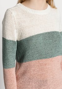 Pull en maille avec un col rond. Présente des rayures horizontales en crème, vert et rose. Tissu en mailles texturé et lâche.