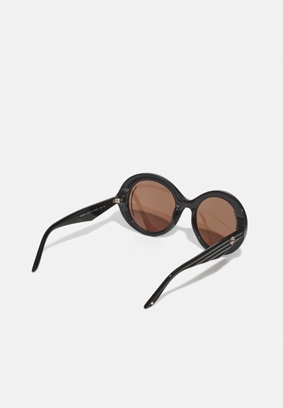 Round sunglasses with glossy black frames and brown tinted lenses. Features sleek black arms with subtle branding detail near the hinge.