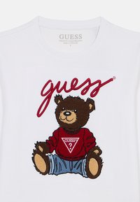 Λευκό μπλουζάκι με κέντημα καφέ αρκουδάκι που φοράει κόκκινο πουλόβερ με τριγωνικό λογότυπο "GUESS?". Κανονική εφαρμογή.