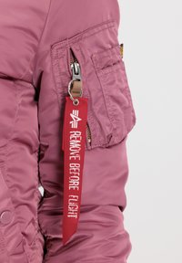 Veste en nylon rose avec une poche latérale, dotée d'une étiquette en tissu rouge sur laquelle est inscrit "ENLEVER AVANT LE VOL" en blanc. Texture lisse, design moderne.