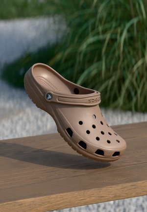 Lysebrun Crocs-klods med ventilationshuller og hælrem, placeret på en træoverflade med en sløret baggrund af udendørs grønne planter.