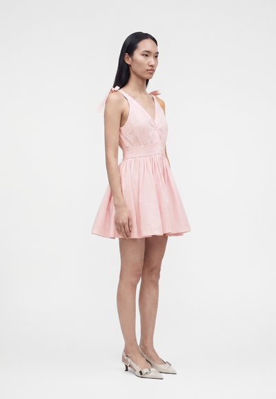 Γυναίκα που φοράει ροζ αμάνικο φόρεμα με δεμένες τιράντες στους ώμους και σφιχτή μέση, φοράει μπεζ χαμηλά παπούτσια, μπροστά σε λευκό φόντο.