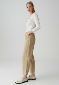 Pantaloni in corda beige chiaro con una vestibilità rilassata, dotati di tasche laterali e un dettaglio di cucitura. Abbinati a una maglietta bianca a maniche lunghe e mocassini.
