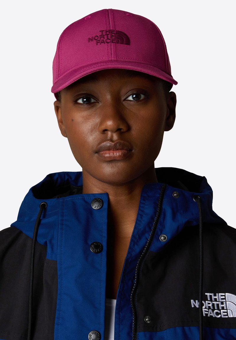 Justerbar rosa keps med broderad logotyp, tillverkad av tyg. Bärs av en person i en blå och svart jacka med knappar och dragsnören.