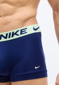 Κοντινό πλάνο ανδρικού κορμού που φοράει σκούρο μπλε μποξεράκια Nike με ανοιχτό πράσινο λάστιχο μέσης που φέρει το λογότυπο και το σήμα της Nike.